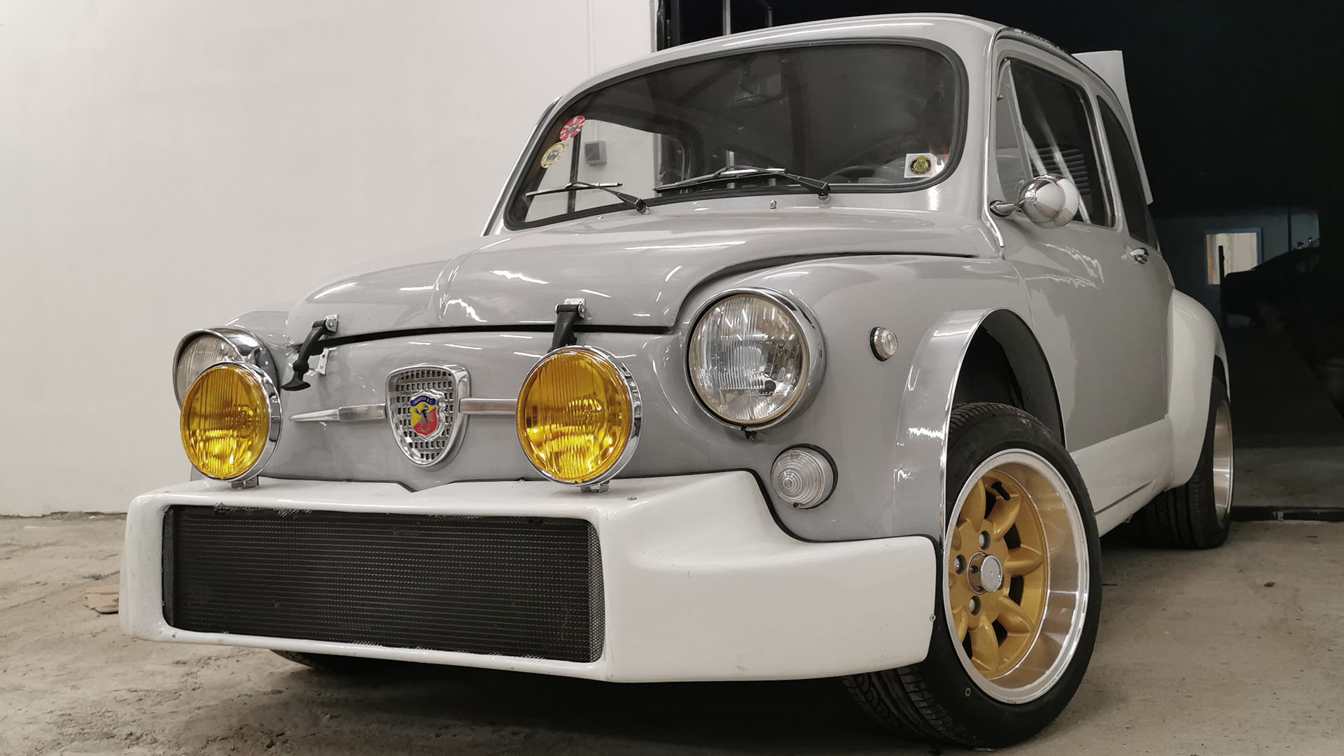 Grijze Abarth 850 Nürburgring klassieke raceauto vooraanzicht met gele verstralers, witte voorbumper met ingebouwde radiateur en gouden Minilite velgen.