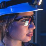 Vrouwelijke ingenieur met slimme veiligheidshelm, hoofdlamp en veiligheidsbril die digitale gegevens bekijkt in een industriële omgeving.